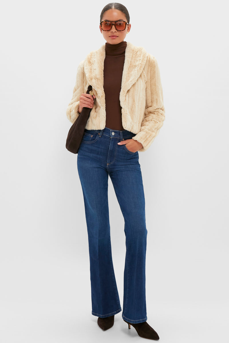 A person wearing sunglasses, a L'AGENCE cream faux fur jacket with a luxe mink look, brown turtleneck, blue flared jeans, dark brown boots, and carrying a brown shoulder bag.