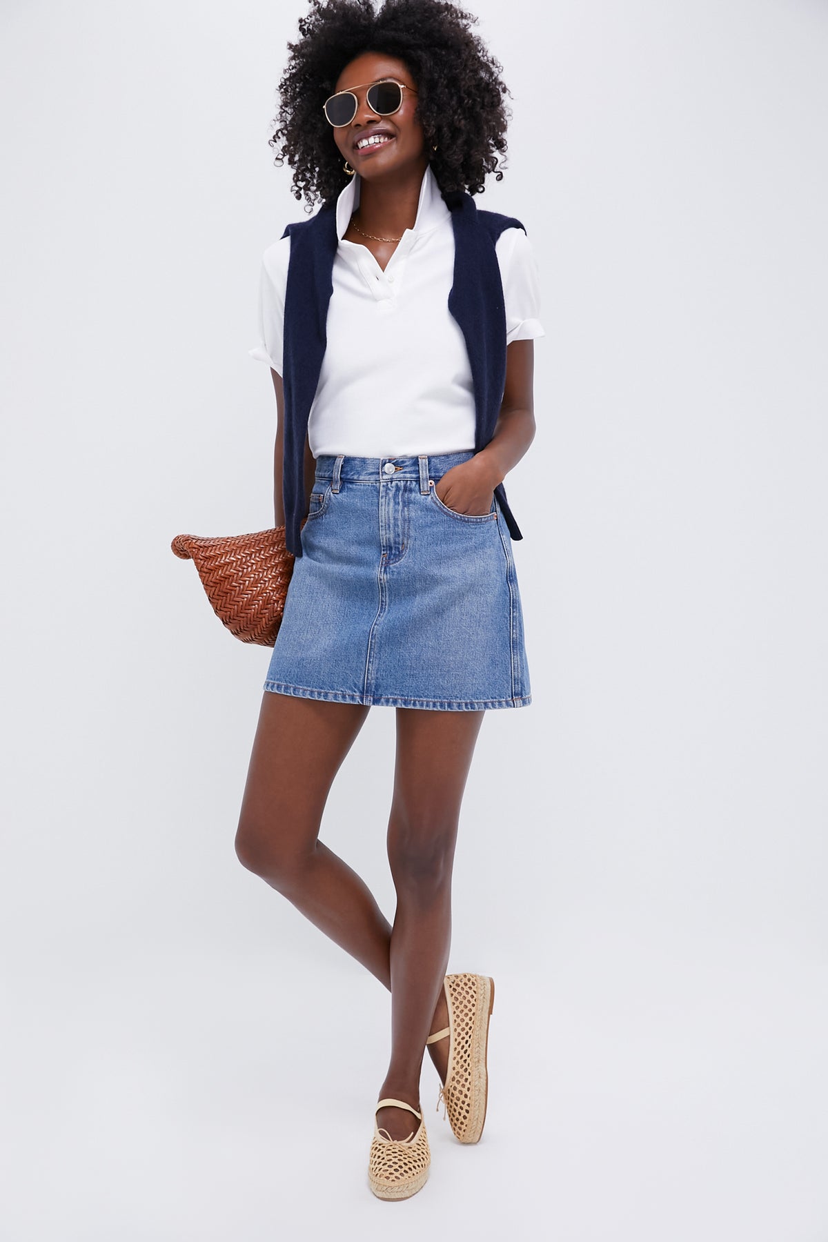 A woman in Tuckernuck sunglasses, a white textured pique polo, a navy sweater over her shoulders, a denim skirt, woven shoes, and holding a brown bag showcases relaxed fit style with effortless chic.