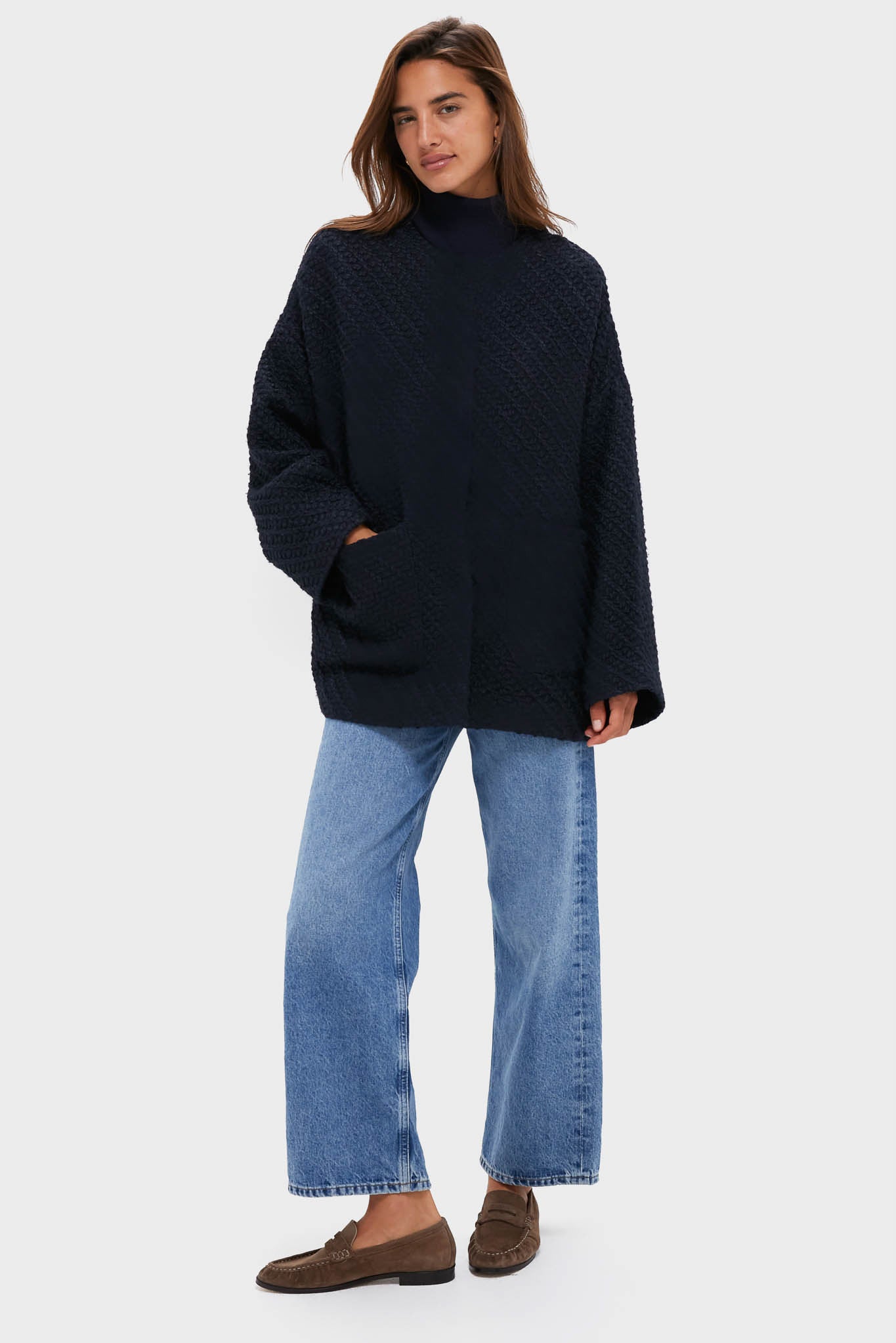 A woman models a Tuckernuck textured black oversized sweater, light blue wide-leg jeans, brown loafers, and a relaxed fit coat against a plain light background.
