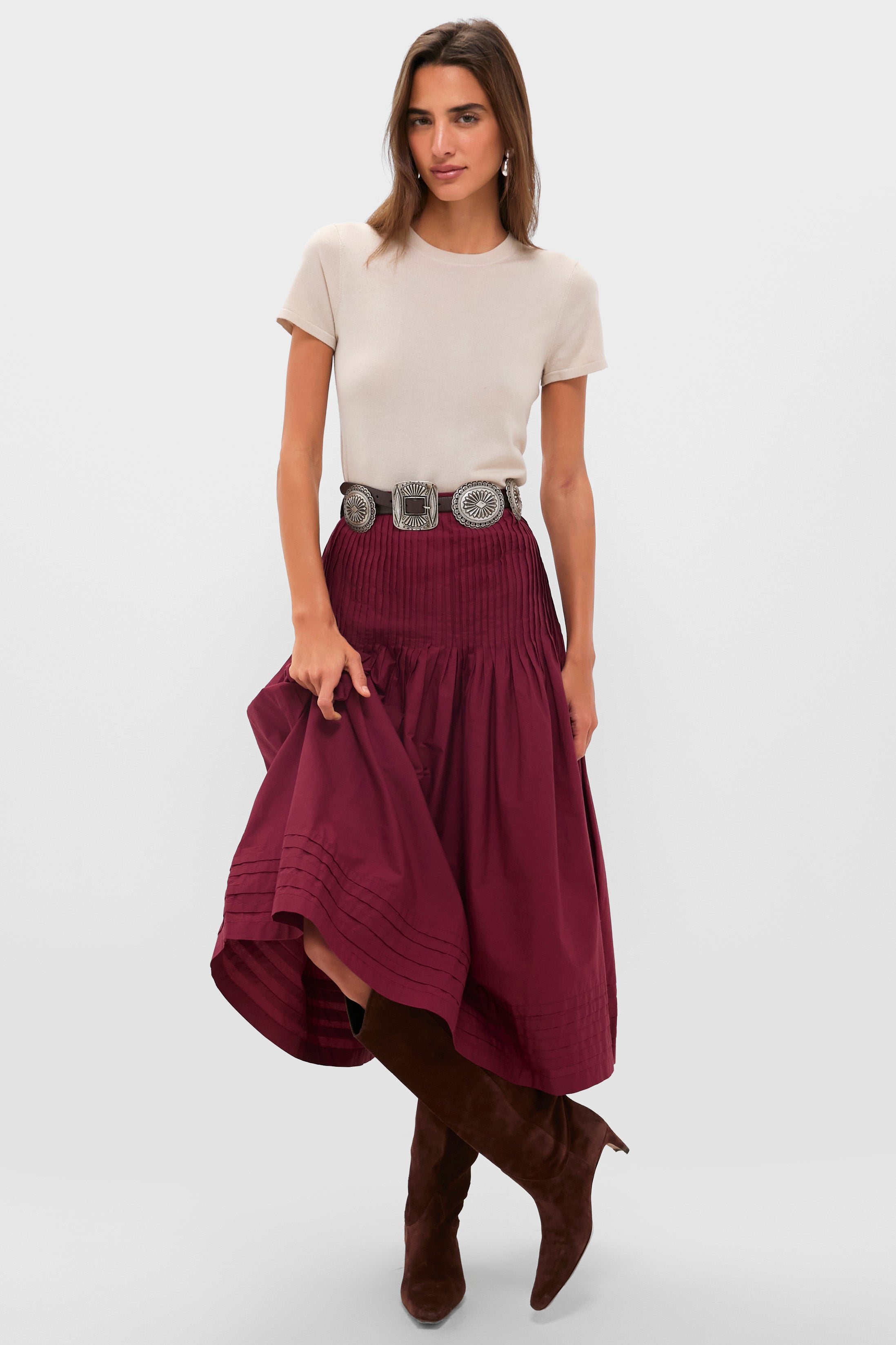 A woman stands against a plain light background wearing a beige short-sleeve top, a pleated Ciao Lucia cotton poplin skirt, a wide silver belt, and brown knee-high boots.