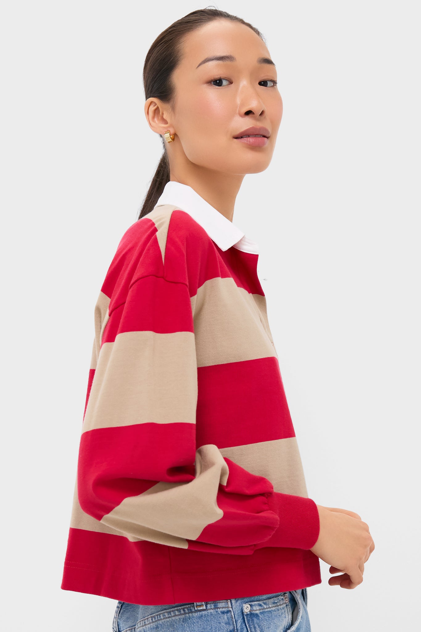 A woman in a boxy, loose-fit red and beige striped Tuckernuck polo with a white collar and blue jeans stands against a plain light background.