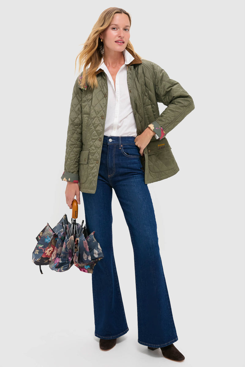 A woman stands against a plain background wearing a green quilted Barbour x Tuckernuck jacket, white shirt, blue flared jeans, brown shoes, and holding a closed Barbour umbrella with a floral pattern.