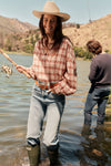 Woman fishing in a lake in a straw hat, plaid shirt, jeans, and boots. 