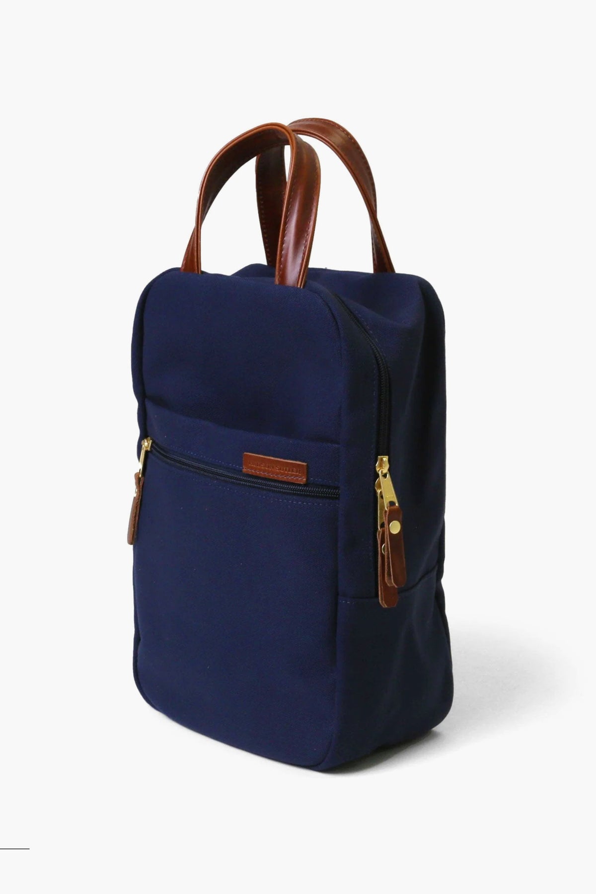 A navy blue rectangular Hudson Sutler backpack with brown leather handles, gold zippers, and a small exterior zip pocket is displayed against a plain white background.
