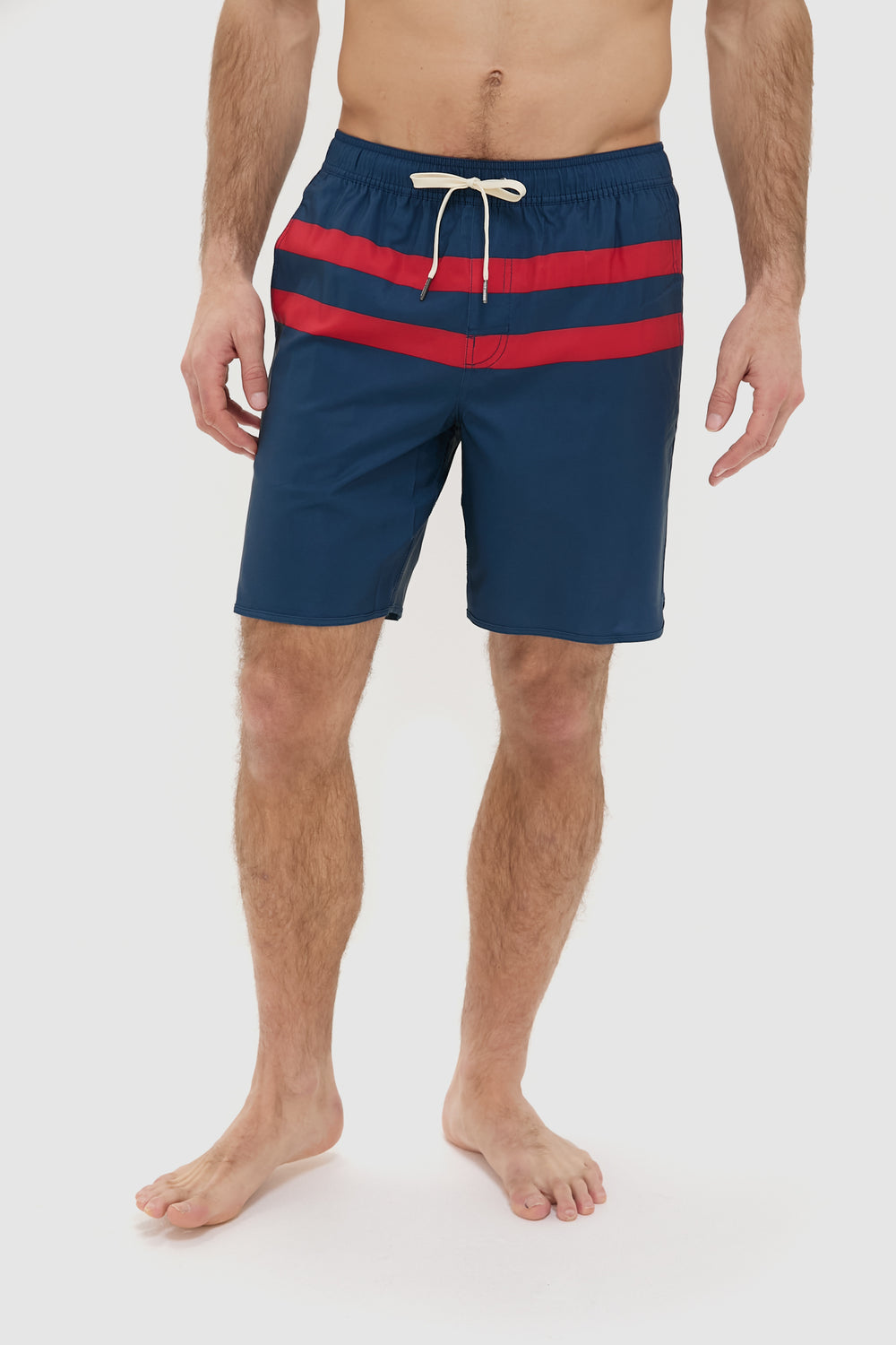 A man wears Fair Harbor navy performance shorts with two red stripes and a white drawstring, standing barefoot. Made from upcycled plastic bottles, these quick-drying shorts are ideal for an active lifestyle.