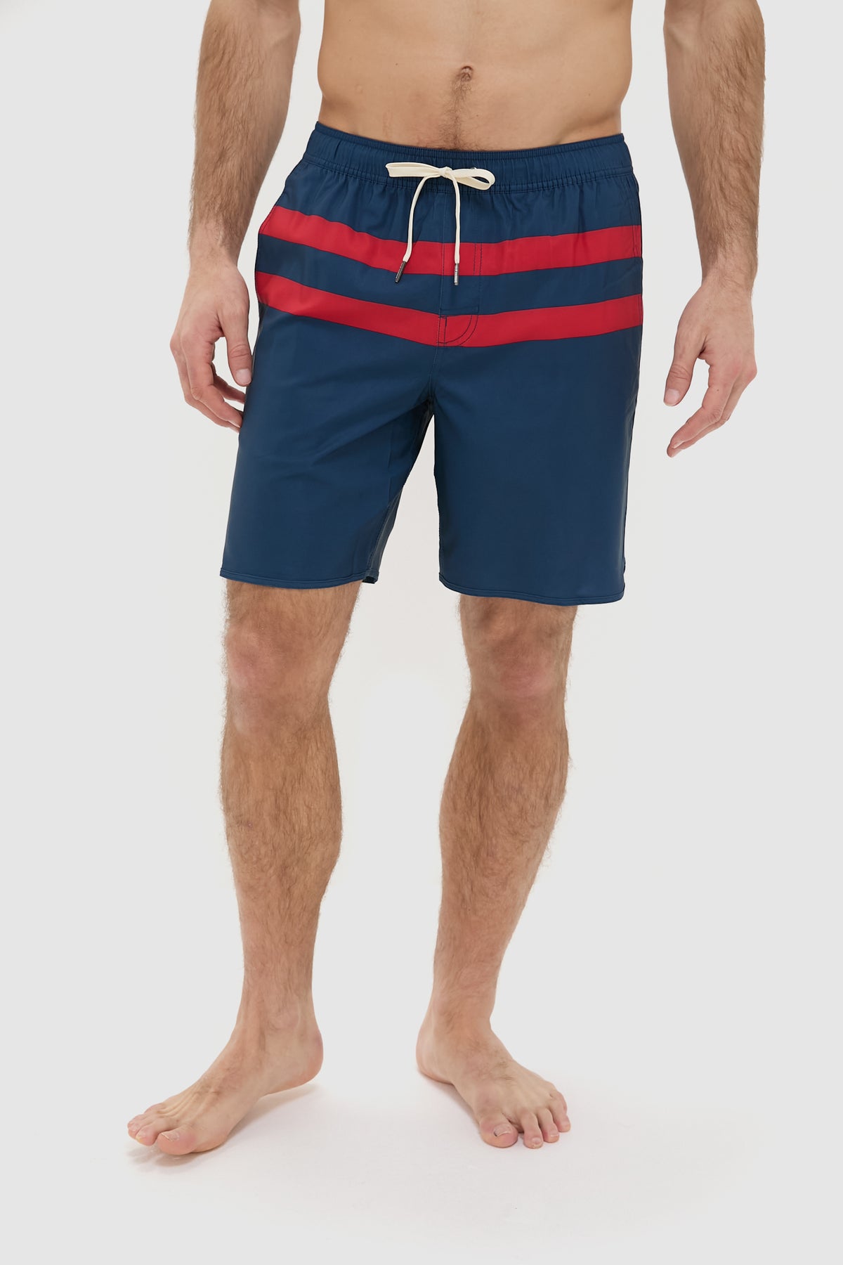 A man wears Fair Harbor navy performance shorts with two red stripes and a white drawstring, standing barefoot. Made from upcycled plastic bottles, these quick-drying shorts are ideal for an active lifestyle.