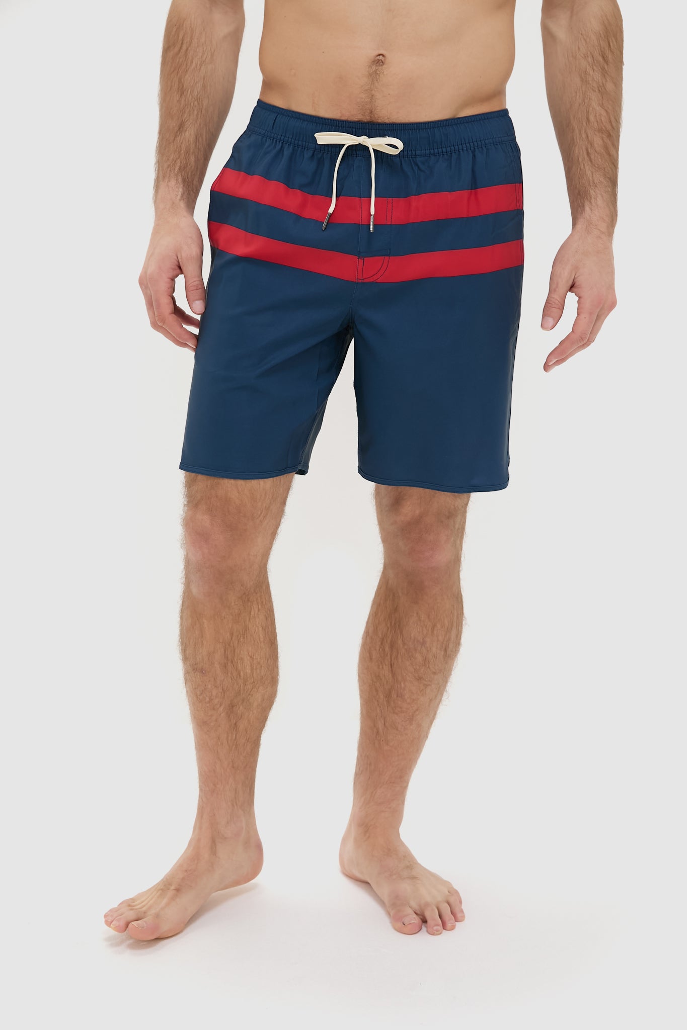A man wears Fair Harbor navy performance shorts with two red stripes and a white drawstring, standing barefoot. Made from upcycled plastic bottles, these quick-drying shorts are ideal for an active lifestyle.