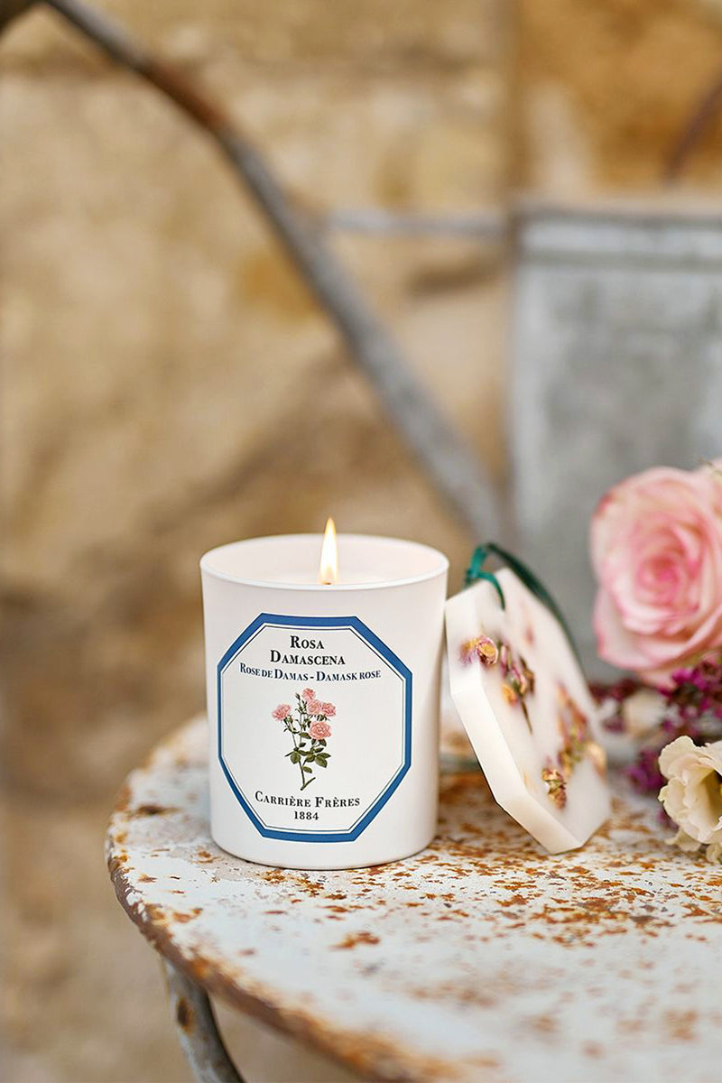 Damask Rose Candle by Carrière Frères on a rustic table, paired with a wax lid adorned with rosebuds. Classic, high-end fragrance, offering effortless elegance and fun luxury.