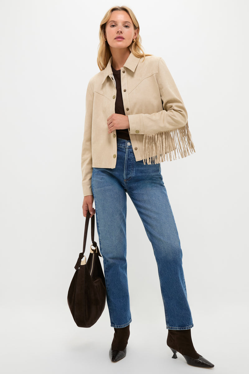A woman poses against a white background in a Hyacinth House western-chic faux suede fringe jacket, dark brown top, blue jeans, pointed black boots, and holding a large brown handbag.