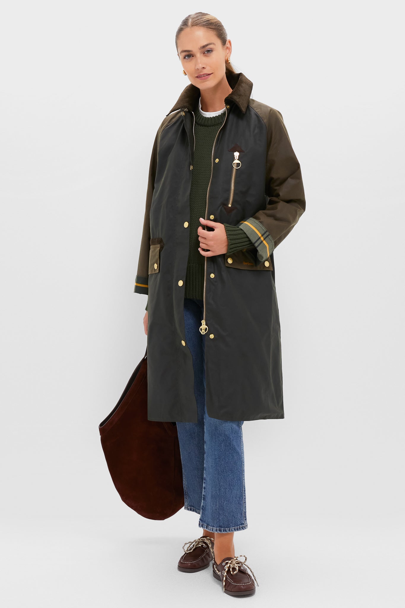 A woman wears a Barbour coat over a green sweater, blue jeans, and brown shoes, holding a large brown bag against a plain light background.