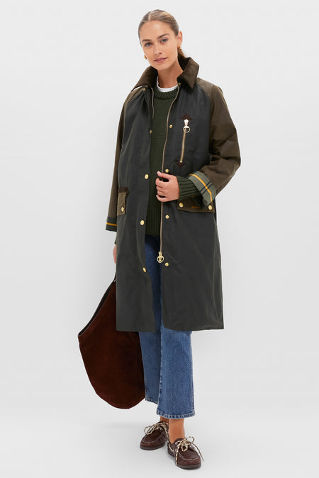 A woman wears a Barbour coat over a green sweater, blue jeans, and brown shoes, holding a large brown bag against a plain light background.