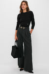 A woman wears a black Tuckernuck crewneck top, dark wide-leg trousers, black belt, and heels, holding a black handbag against a plain background.