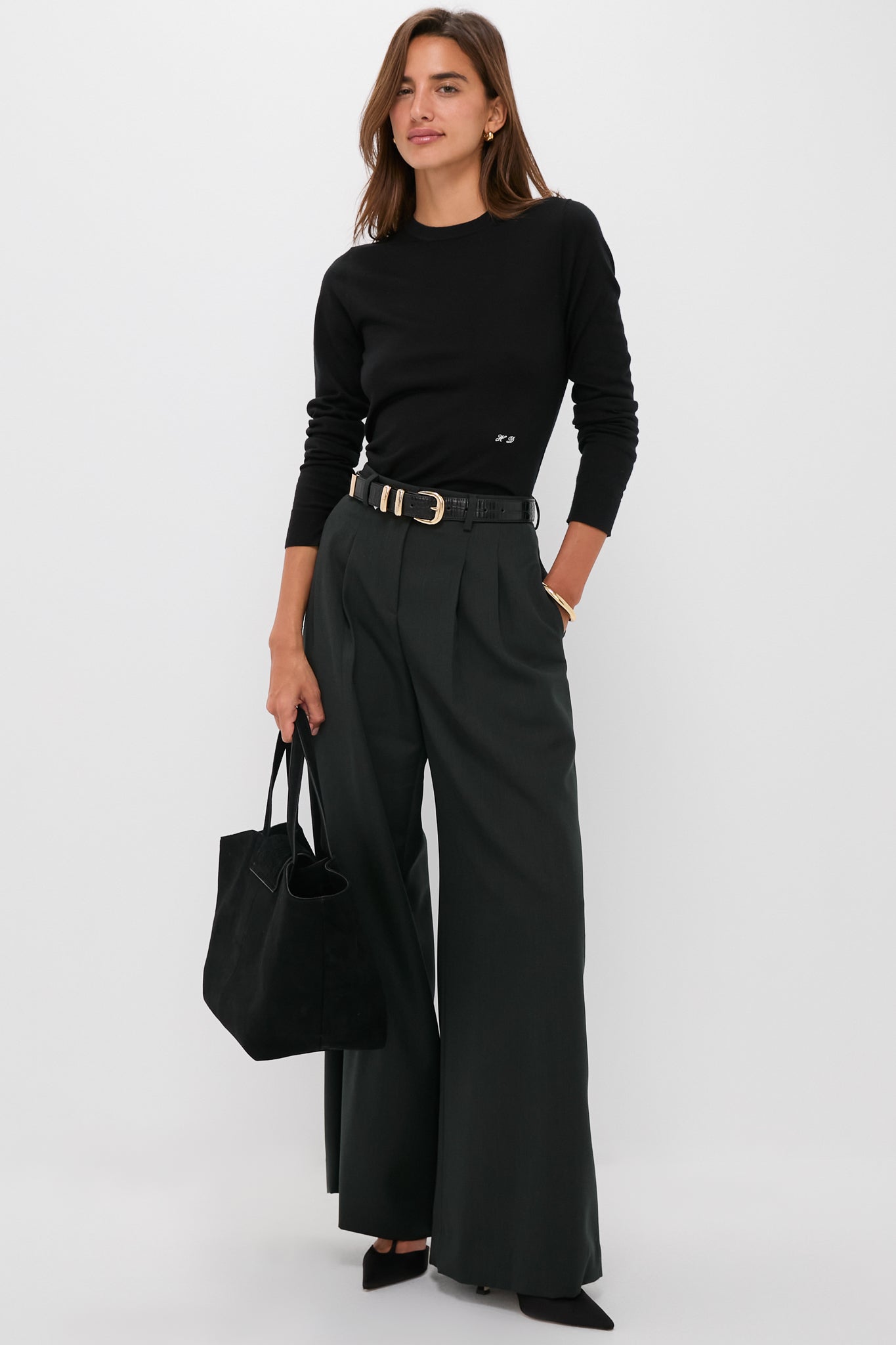 A woman wears a black Tuckernuck crewneck top, dark wide-leg trousers, black belt, and heels, holding a black handbag against a plain background.