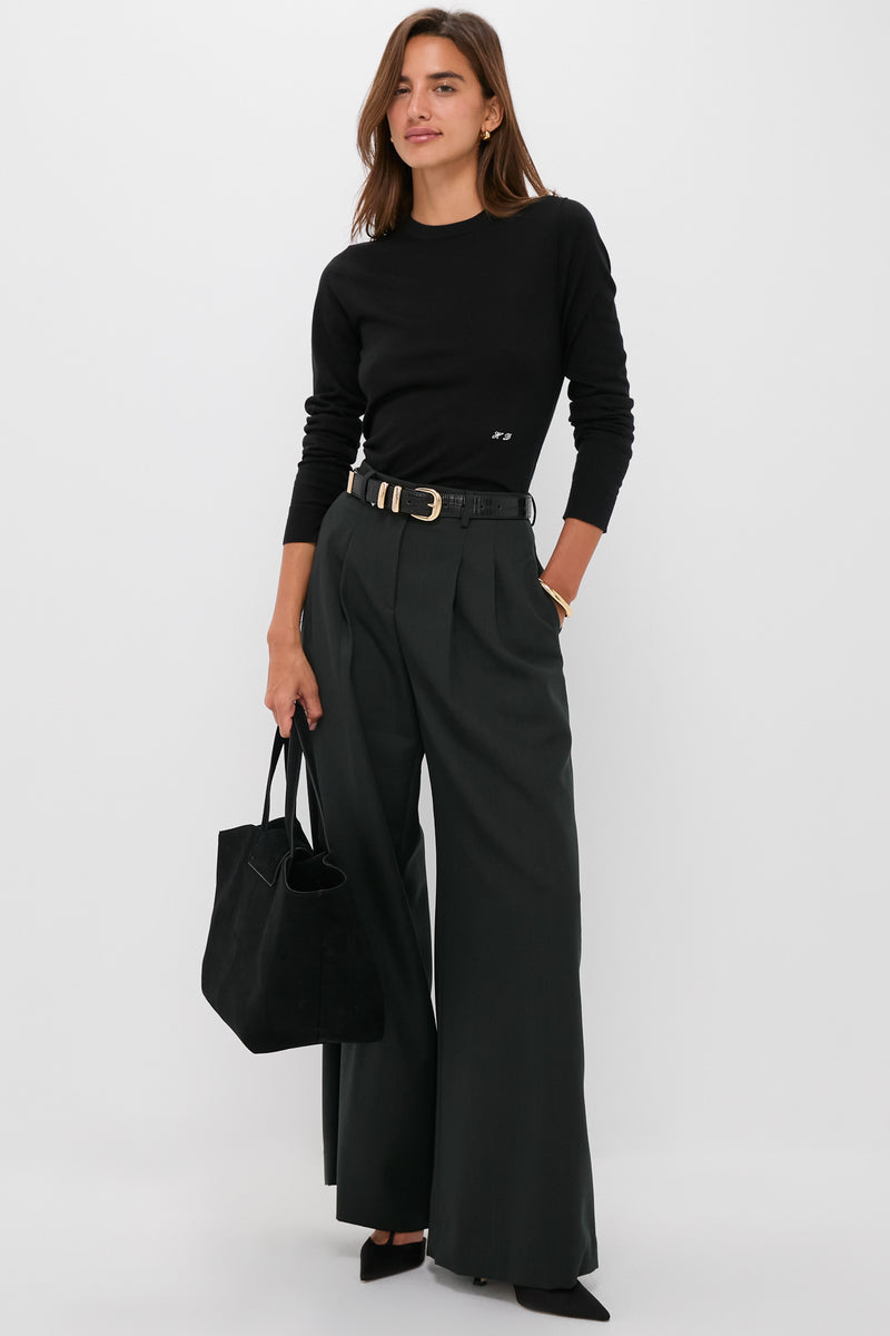 A woman wears a black Tuckernuck crewneck top, dark wide-leg trousers, black belt, and heels, holding a black handbag against a plain background.