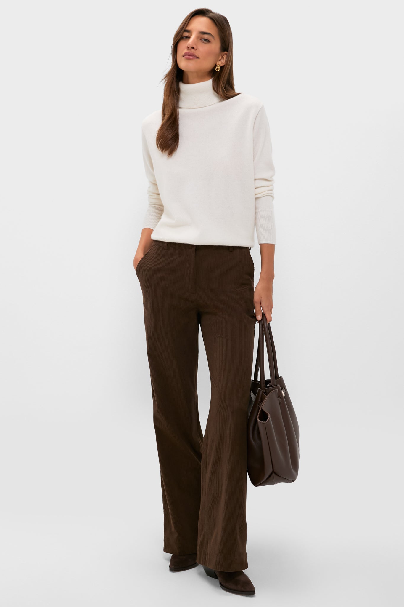 A woman stands against a plain light background wearing a Tuckernuck cashmere turtleneck sweater, brown wide-leg pants, dark shoes, and holding a brown tote bag.