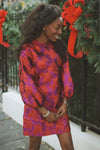 Young woman in a classic, high-end Red & Purple Jacquard Janie Mini Dress with puff sleeves, embodying effortless elegance and fun amidst festive outdoor decor.