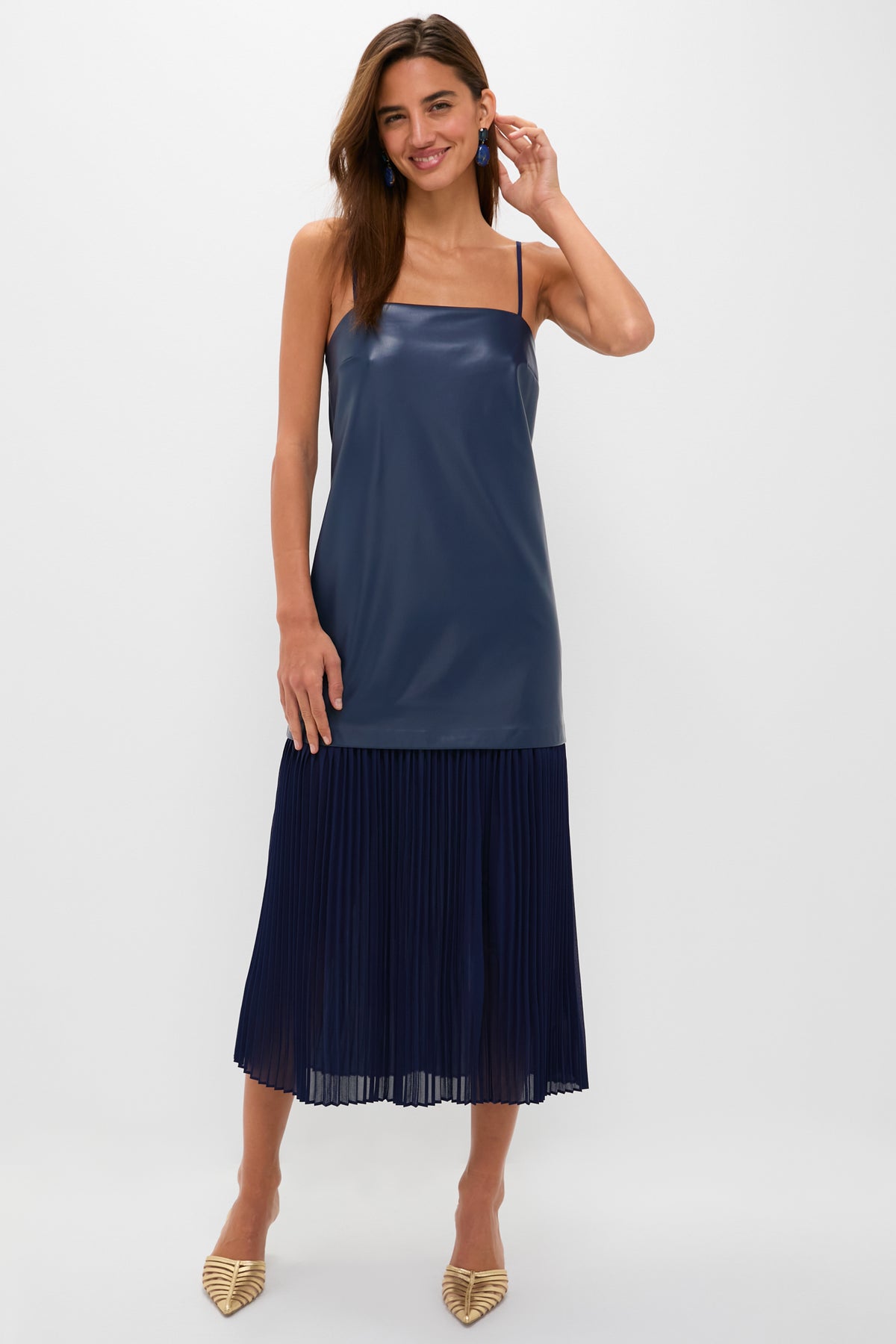 A woman wearing the Hyacinth House Ozzy Midi Dress—a sleeveless navy blue cocktail dress with a shiny top and pleated skirt—paired with pointed-toe beige heels.