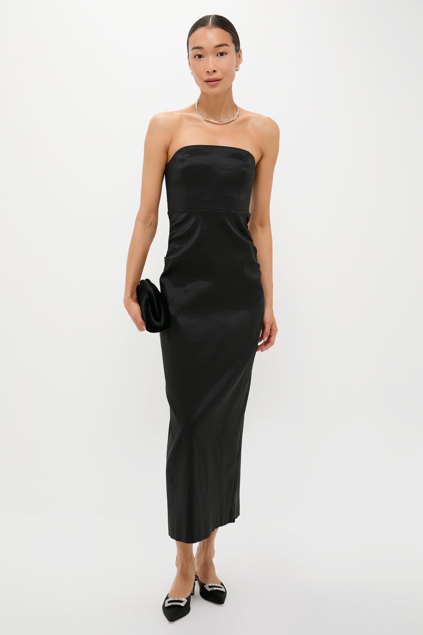 A woman stands against a plain background wearing a strapless black Tuckernuck dress with a sleek sheath silhouette, black pointed shoes, and holding a black clutch.