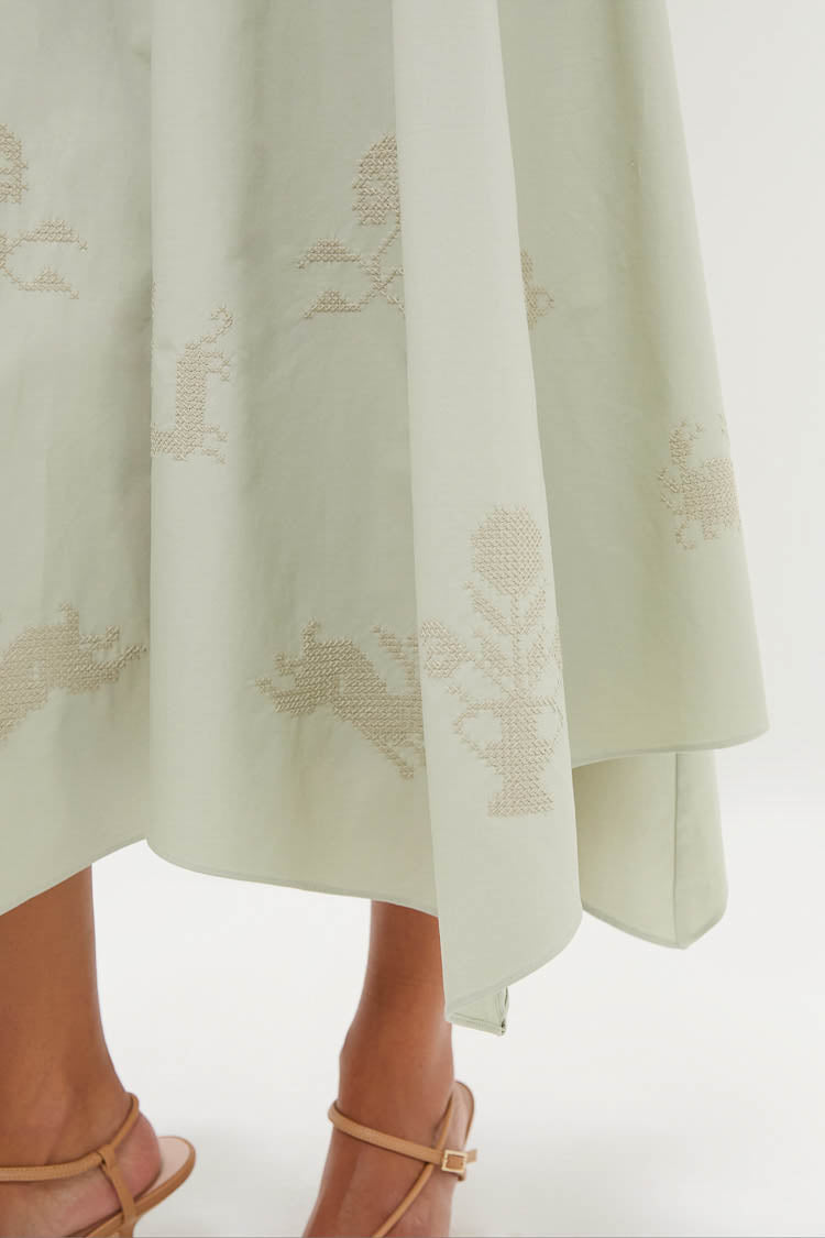 A close-up of a CLEA pistachio Jacqueline dress with subtle embroidered animal and plant motifs, styled as a skirt with tan strappy sandals—a premium womenswear look.