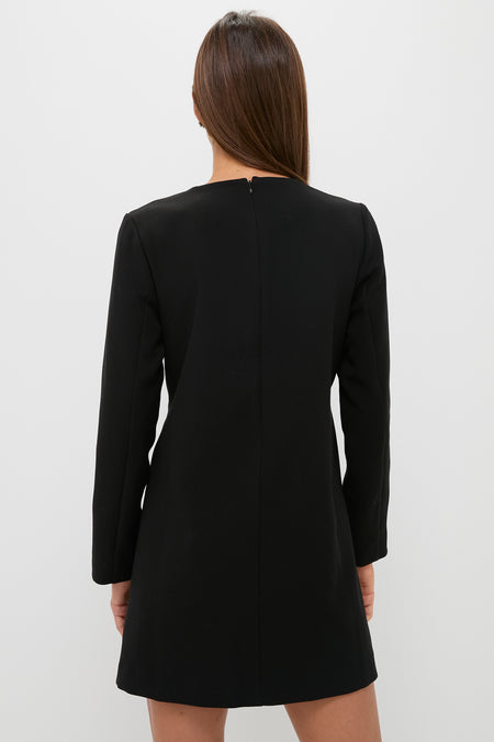 A woman with straight brown hair stands with her back to the camera, modeling a Tuckernuck long-sleeved black crepe bow coat dress against a plain white background.