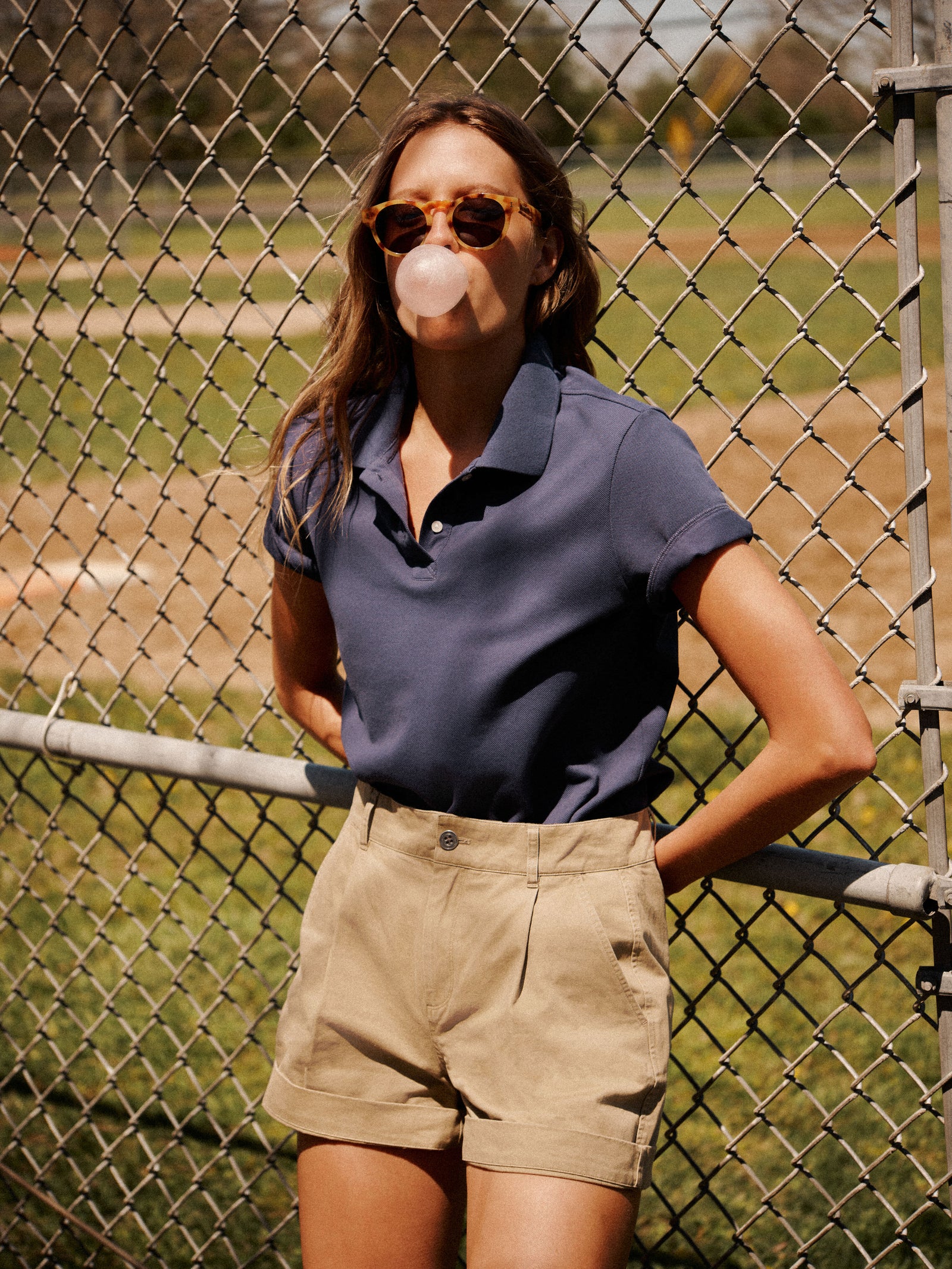 A person in Tuckernuck sunglasses, a blue Holmes Polo in textured pique fabric, and khaki shorts stands by a chain-link fence, blowing a gum bubble.