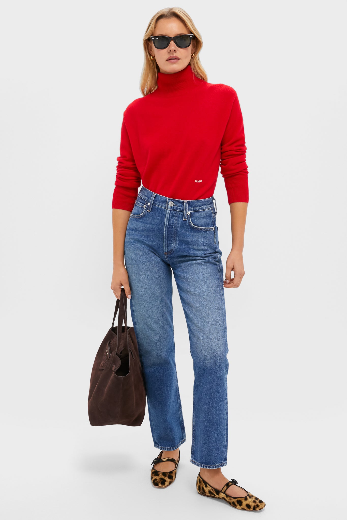 A woman in a red Tuckernuck cashmere turtleneck sweater, blue jeans, leopard print flats, and sunglasses holds a brown bag against a plain background.