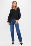 A woman wears a relaxed fit black long-sleeve top by Tuckernuck, blue jeans, black pointed heels, and holds a black clutch bag against a neutral background.