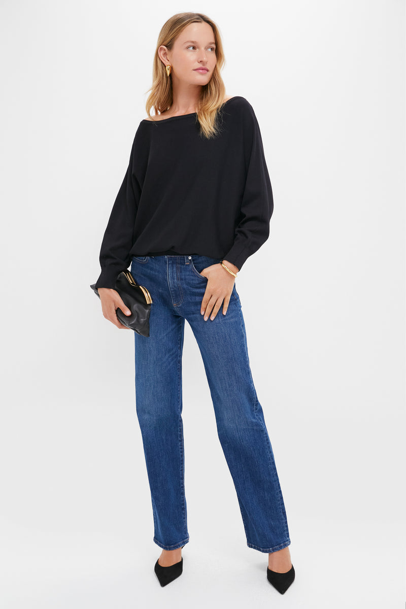 A woman wears a relaxed fit black long-sleeve top by Tuckernuck, blue jeans, black pointed heels, and holds a black clutch bag against a neutral background.