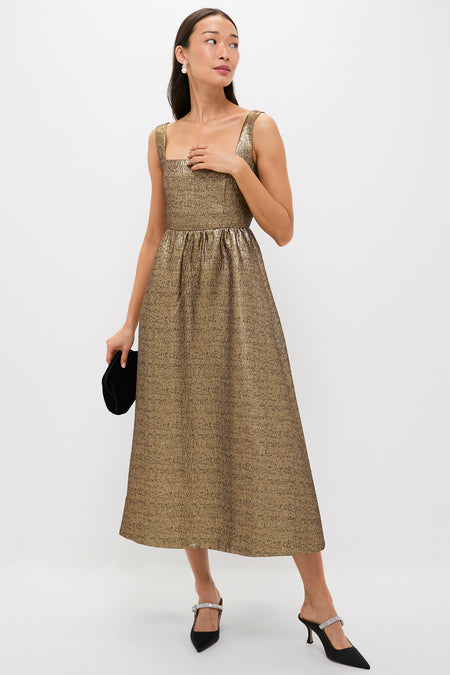 Woman in a metallic gold dress showcases Black Satin Mae Heels, featuring a classic mule silhouette and crystal arch strap—a fun, high-end touch for effortless style.