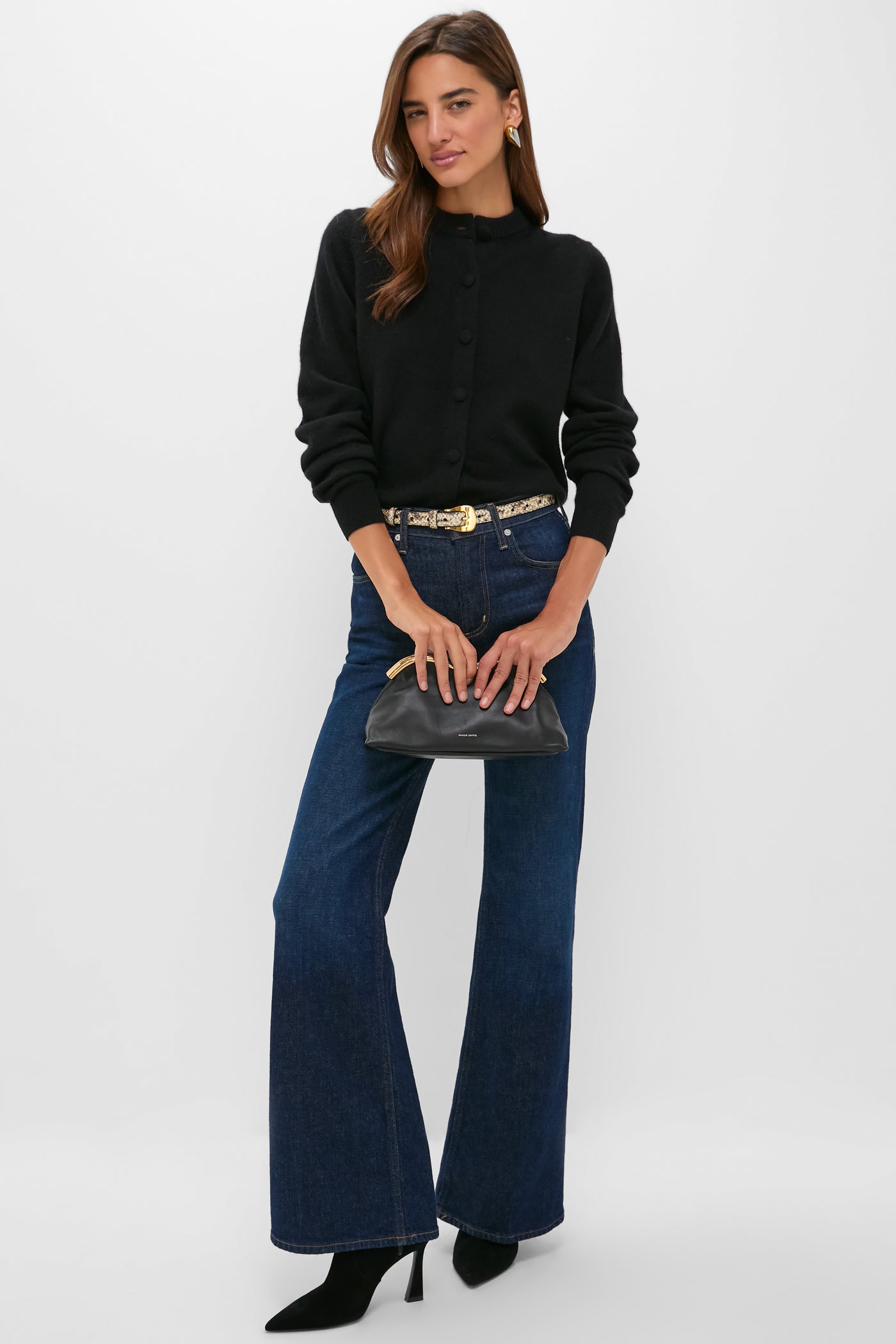 A woman wears a Tuckernuck black cashmere cardigan, blue flare jeans, black heeled boots, and holds a black clutch. Gold belt and earrings complete this timeless fall look.