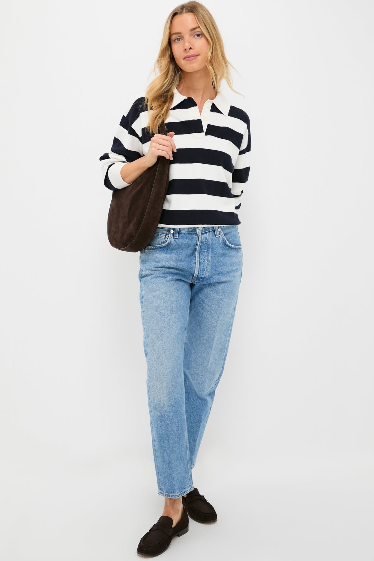 A woman stands against a white background wearing an Alex Mill black-and-white striped polo sweater, light blue jeans, black loafers, and carrying a dark shoulder bag.