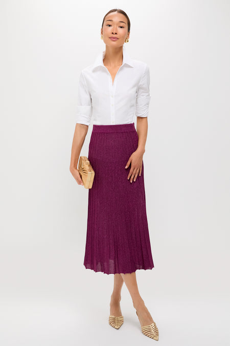 A woman stands against a white background wearing a Tuckernuck white button-up shirt, purple pleated shimmer skirt, nude pointed-toe shoes, and holding a beige clutch.