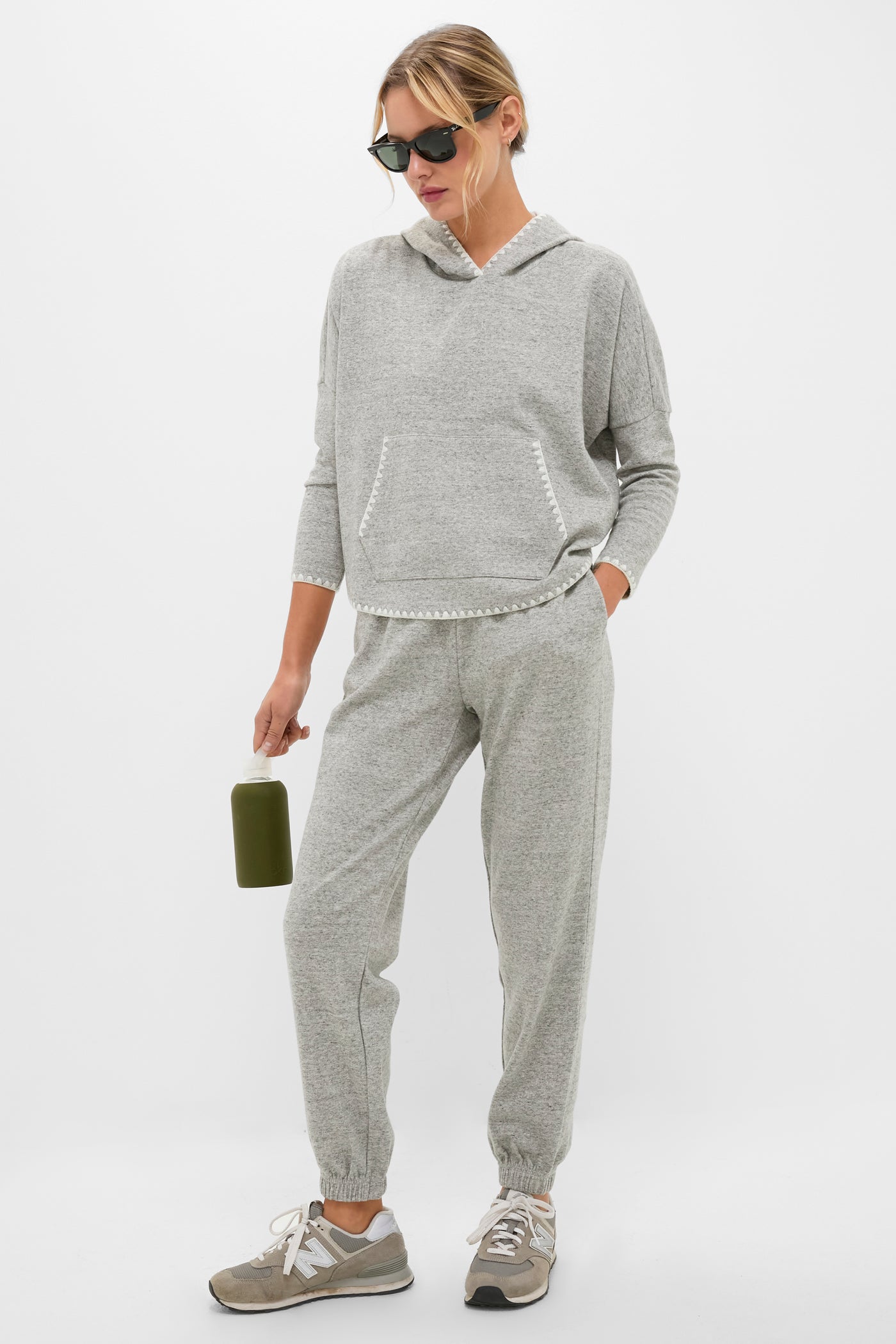 A person wearing a Tnuck Sport light gray hooded sweatshirt with contrast stitching, matching sweatpants, gray sneakers, sunglasses, and holding a green bottle stands against a plain white background.