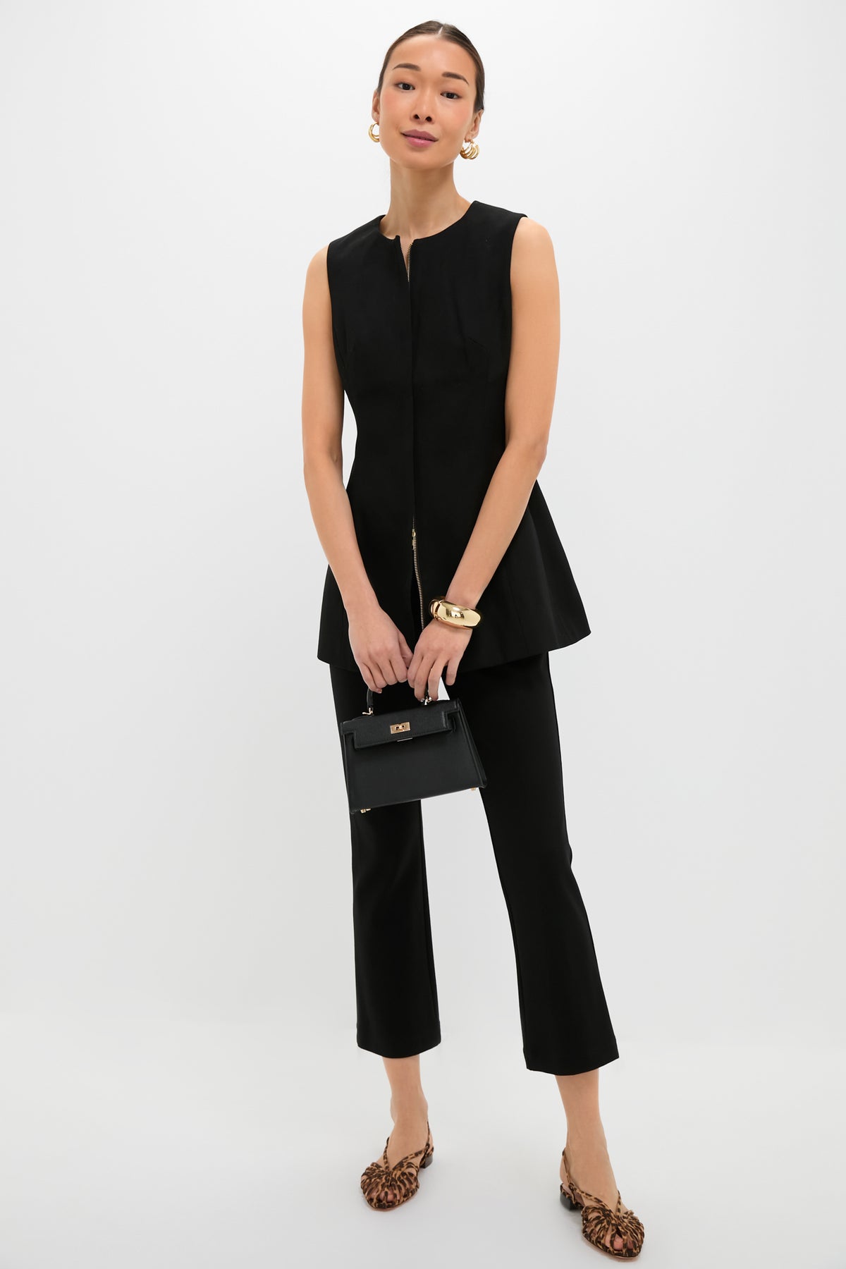 A woman wears a Tuckernuck sleeveless blouse and black cropped pants, carrying a small black handbag. She completes her look with leopard print flats, gold hoop earrings, and bracelets for a polished finish.