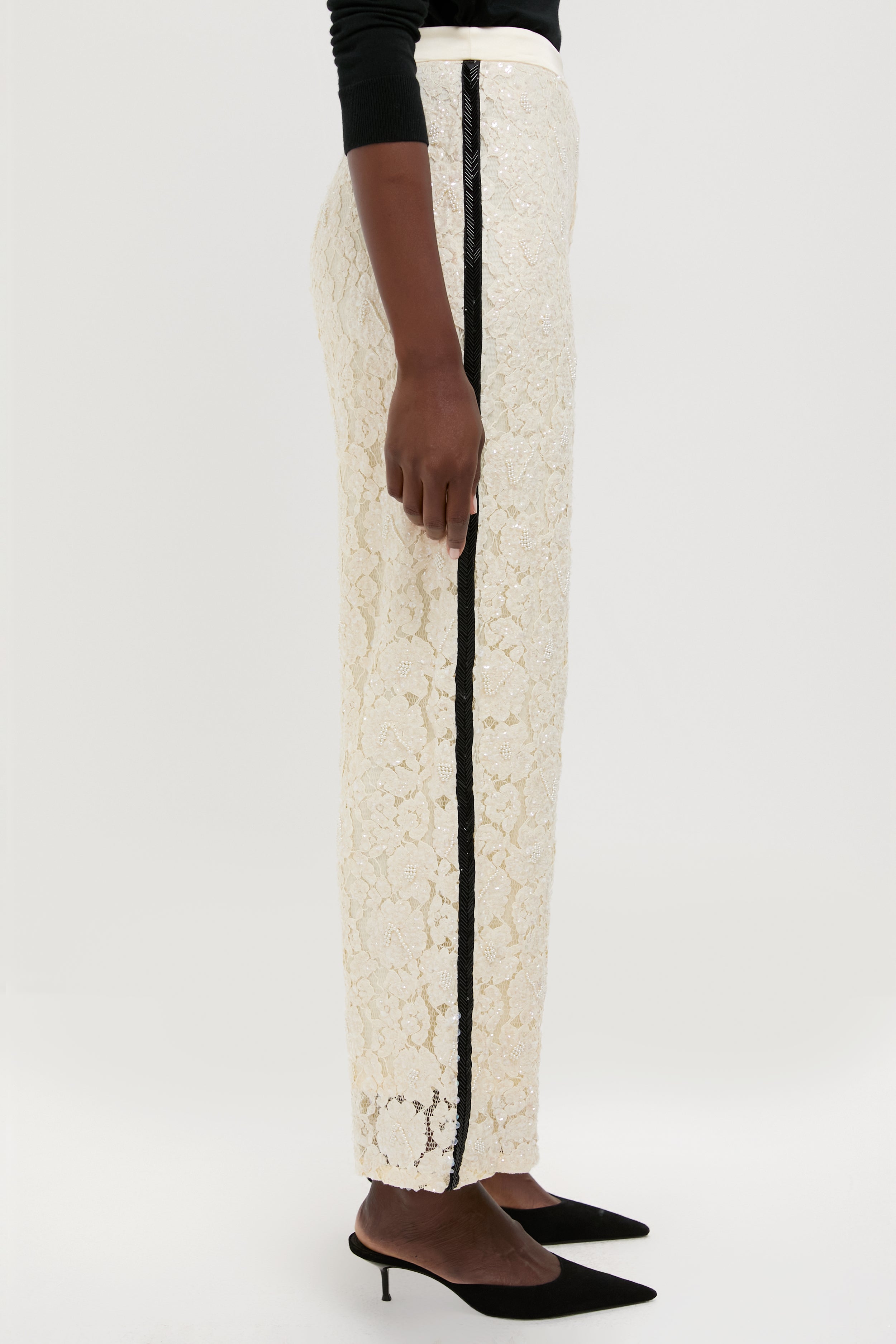 A person in Polo Ralph Lauren floral lace pants with a black vertical stripe, a black long sleeve top, and black pointed-toe low heels, photographed in profile.