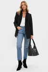 A woman stands against a white background wearing a Tuckernuck Black Structured Olivier Blazer, white top, blue jeans, black heeled boots, and holding a large black handbag.
