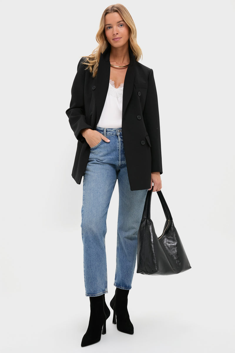 A woman stands against a white background wearing a Tuckernuck Black Structured Olivier Blazer, white top, blue jeans, black heeled boots, and holding a large black handbag.
