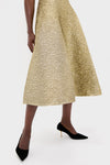 A person wears an Emilia Wickstead gold textured midi skirt embodying sculptural glamour, paired with black pointed-toe heels, standing against a plain white background.