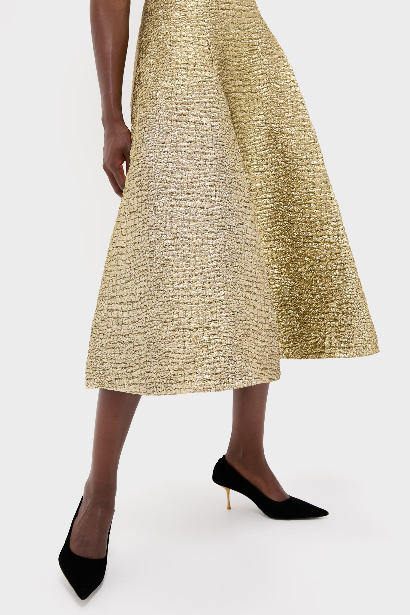 A person wears an Emilia Wickstead gold textured midi skirt embodying sculptural glamour, paired with black pointed-toe heels, standing against a plain white background.