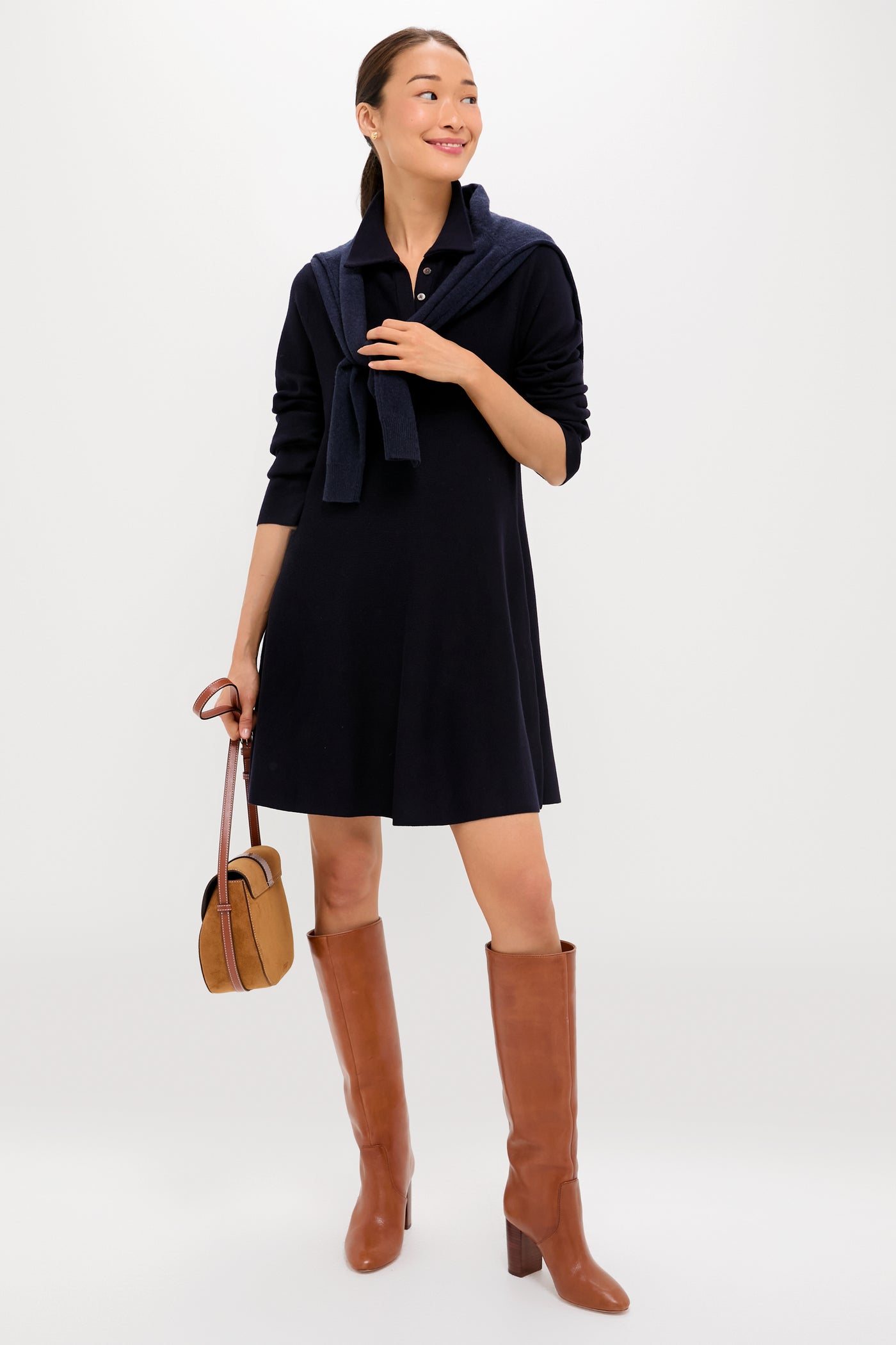 A woman wears a black Tuckernuck Callahan Shirt Dress with a relaxed fit, brown knee-high boots, a navy sweater draped over her shoulders, and holds a brown handbag, standing against a plain background.