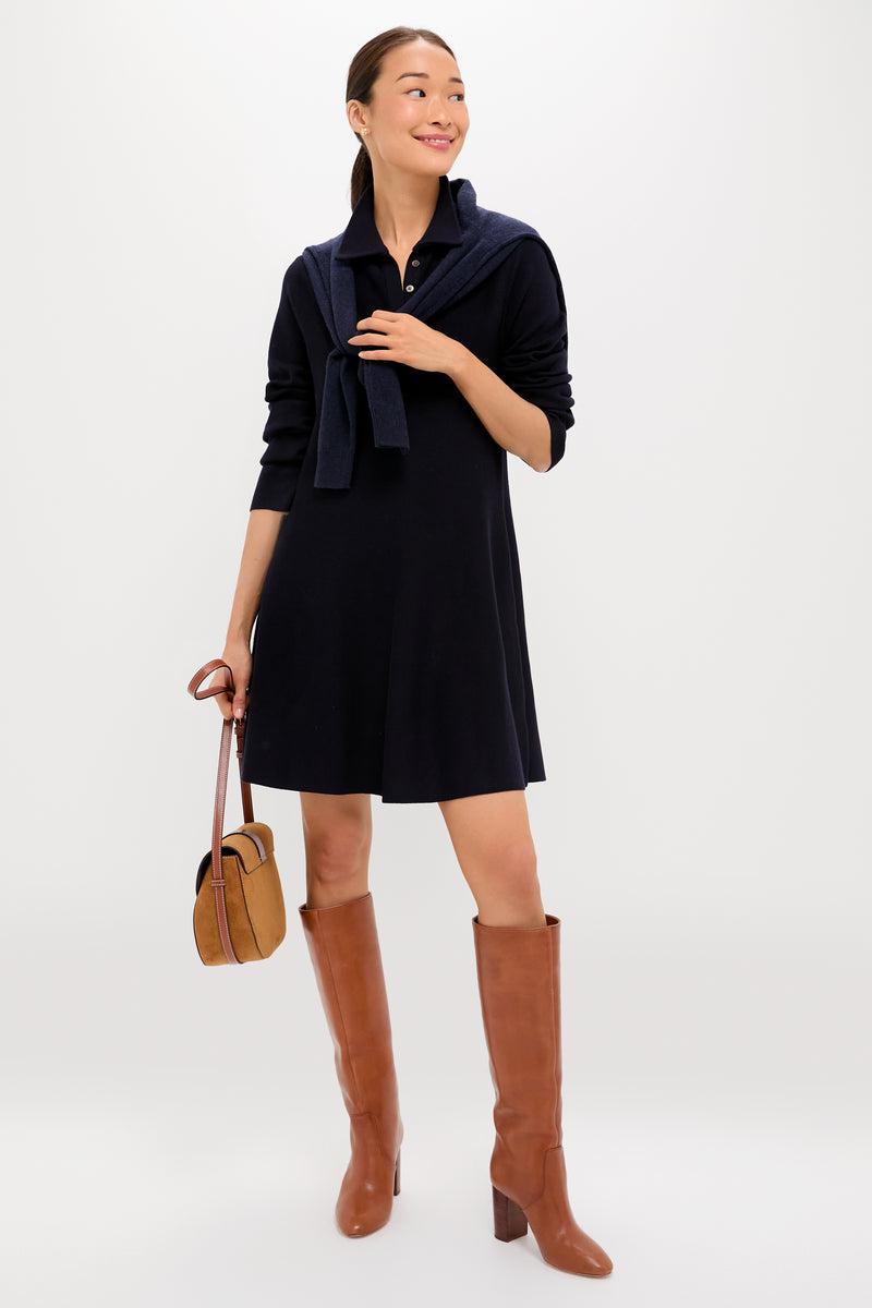 A woman wears a black Tuckernuck Callahan Shirt Dress with a relaxed fit, brown knee-high boots, a navy sweater draped over her shoulders, and holds a brown handbag, standing against a plain background.