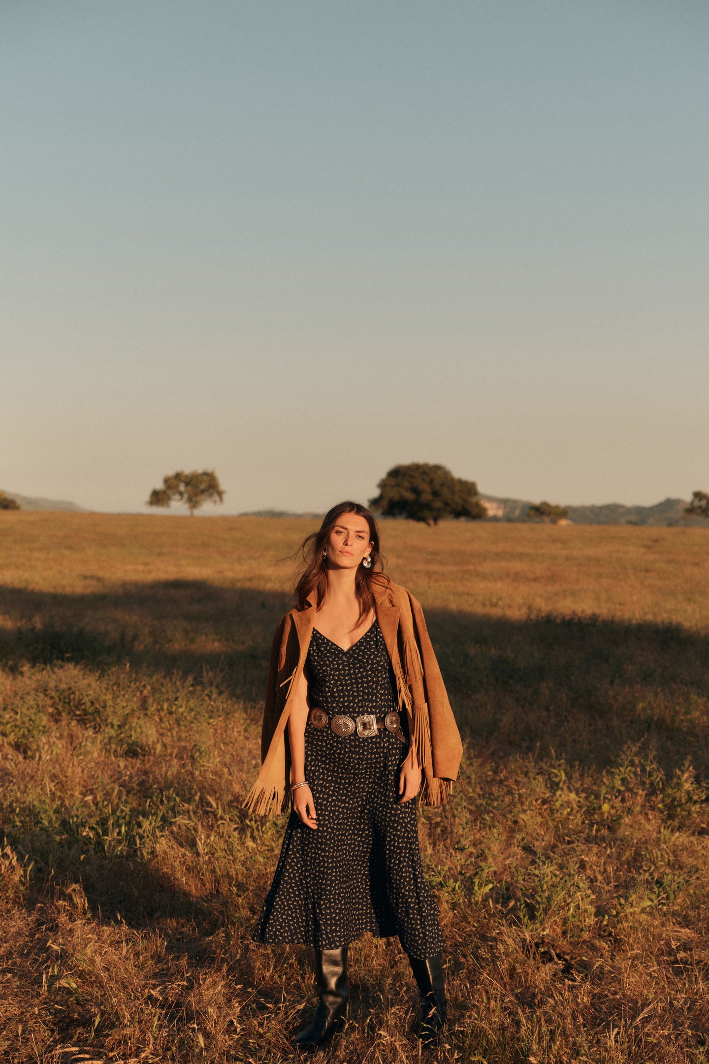 Woman standing outside in a black slip dress with a brown jacket around her shoulders. 