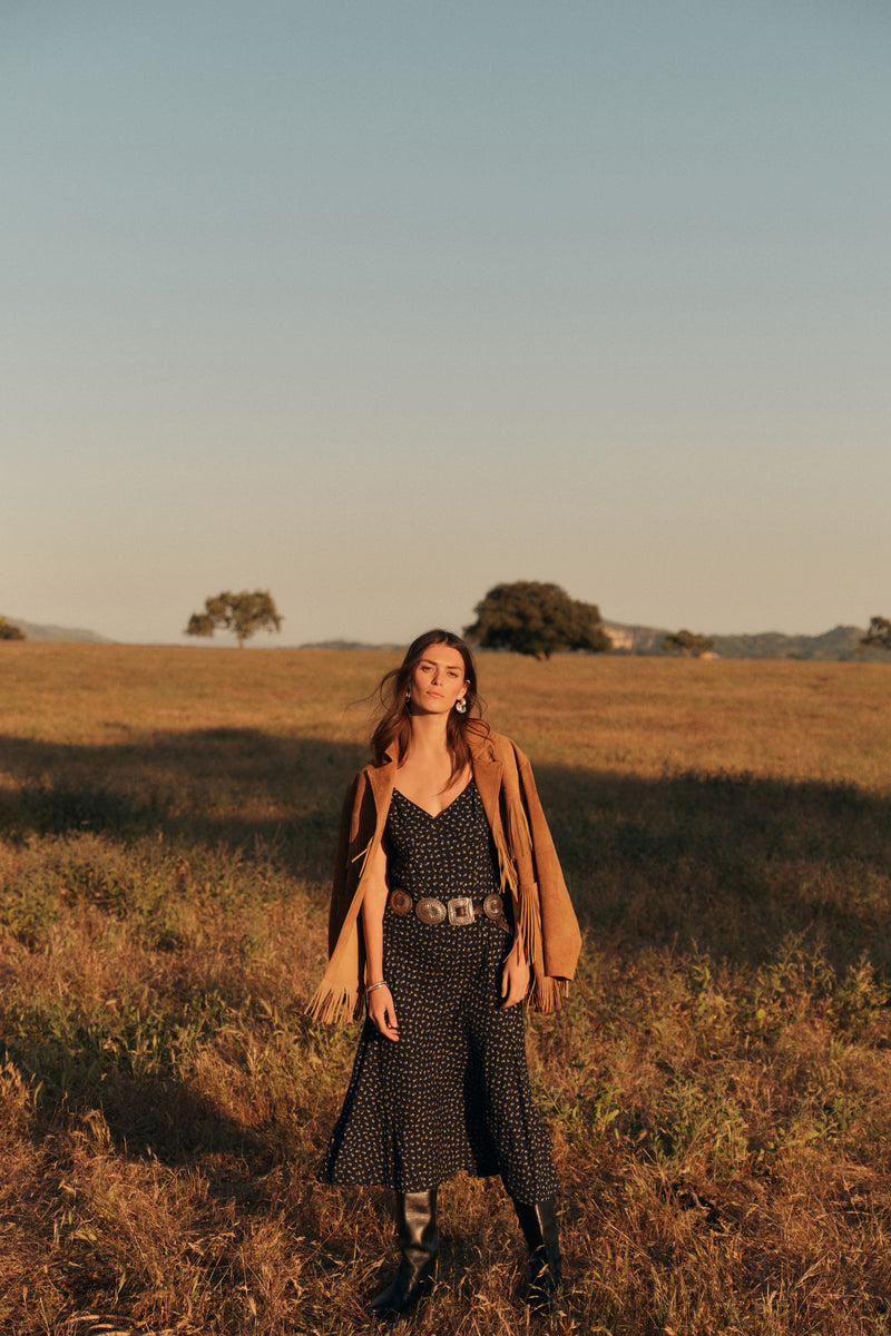 Woman standing outside in a black slip dress with a brown jacket around her shoulders. 