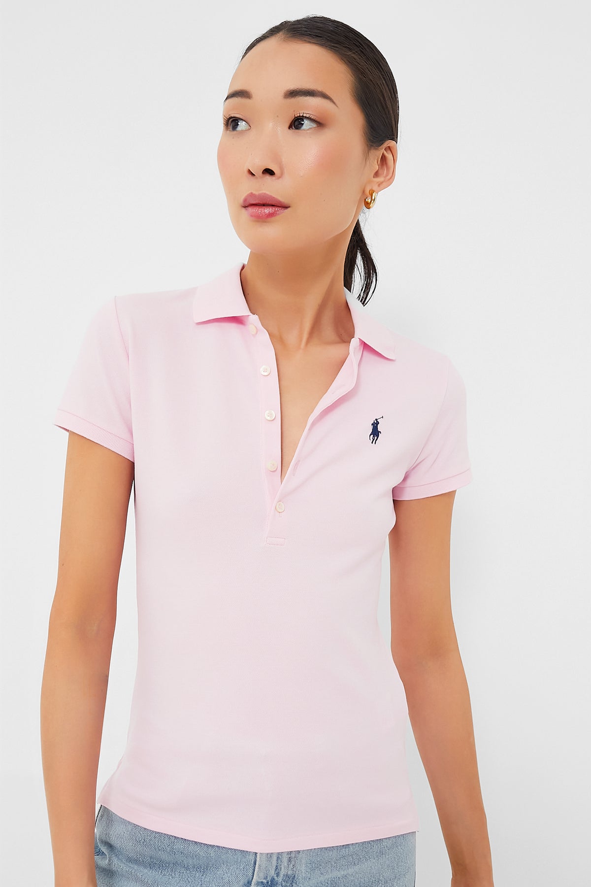 A woman wears a Polo Ralph Lauren slim polo shirt in sunkissed pink with a navy logo, paired with light blue jeans.