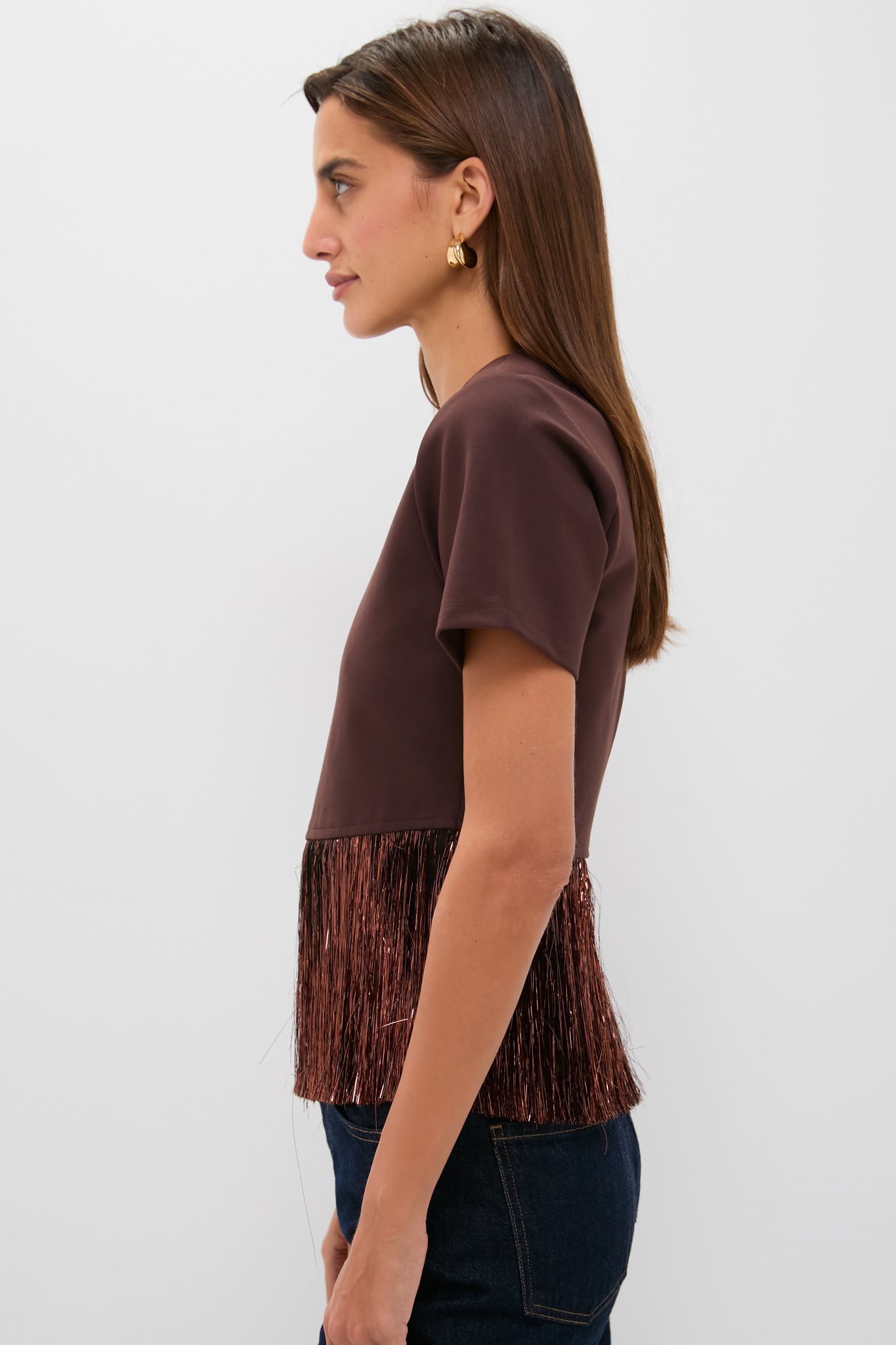 Young woman in profile wearing the Bronze Metallic Fringe Francie Short Sleeve Blouse, showcasing its classic, fun, and high-end fringe detail. Paired effortlessly with dark blue jeans.