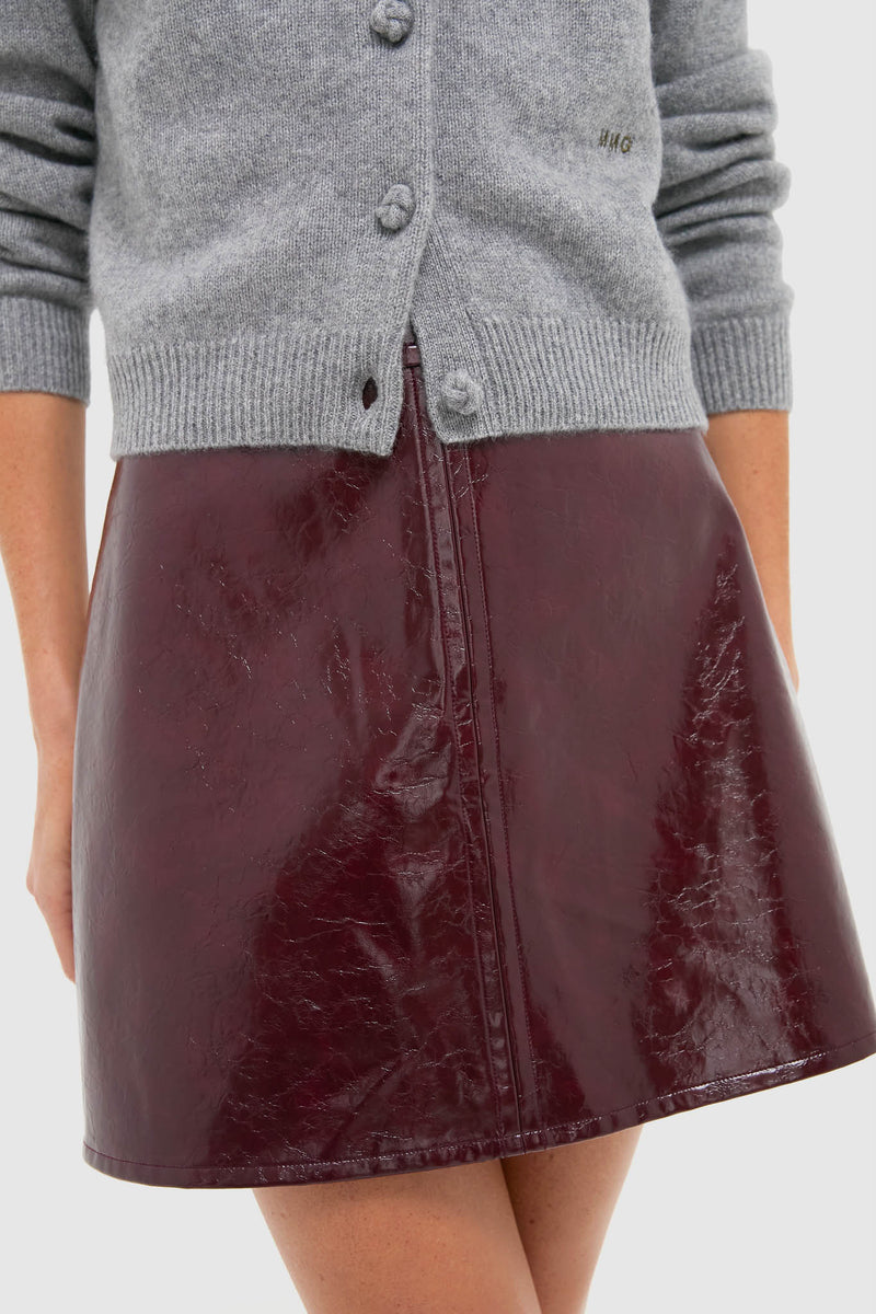 Oxblood Meeghan Mini Skirt in glossy vegan leather, showcasing a classic, effortless style with a front seam, paired with a light-grey knitted cardigan for a high-end, fun look.