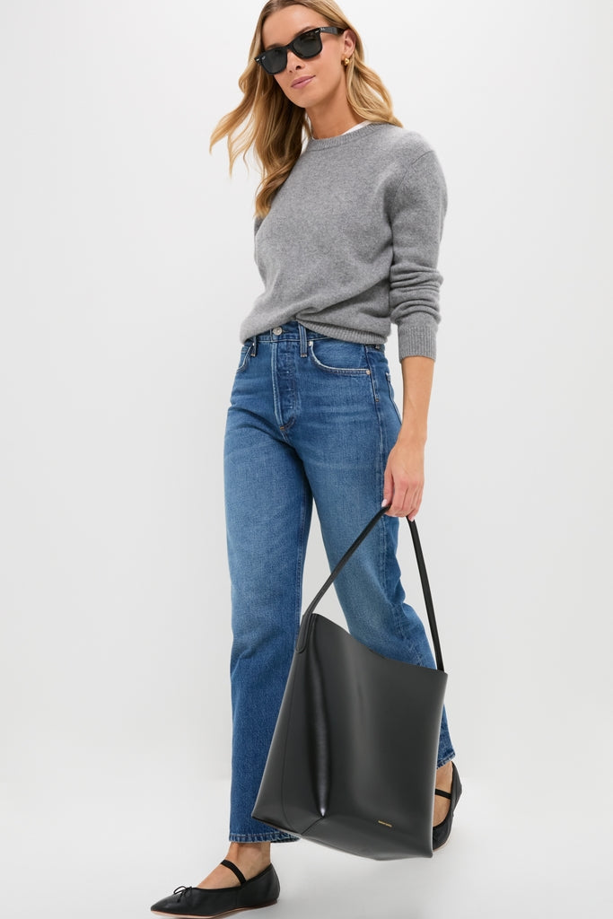 Woman wearing a gray sweater, blue jeans, black flats, and sunglasses holds a Mansur Gavriel luxury tote bag made from Italian spazzolato leather; background is plain white.