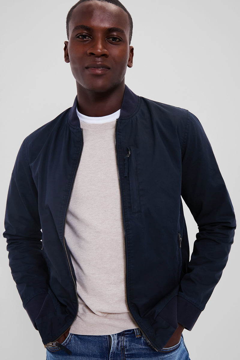 A man stands with his hands in his pockets, wearing a Marine Layer navy water-resistant waxed cotton bomber jacket over a light beige sweater and blue jeans, showcasing a vintage look.