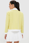 Woman in a pale-yellow Lemongrass Cable Charley Cardigan, featuring a cable-knit design with white trim, paired with a white pleated mini-skirt for an effortless, classic style.