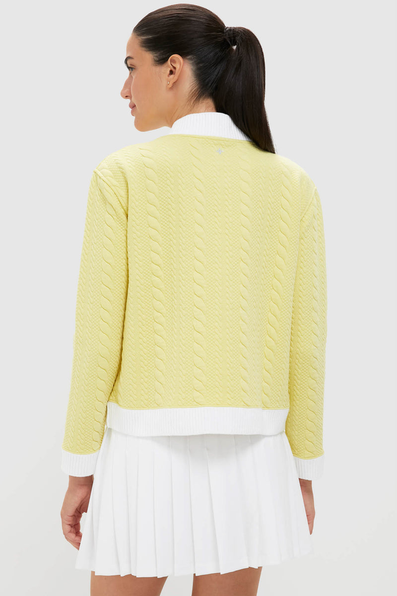 Woman in a pale-yellow Lemongrass Cable Charley Cardigan, featuring a cable-knit design with white trim, paired with a white pleated mini-skirt for an effortless, classic style.